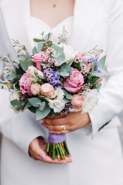 A person in a white outfit holding a fresh bouquet of floral arrangement featuring pink roses, light purple and white flowers, and sprigs of green eucalyptus leaves. The bouquet is wrapped with a purp
