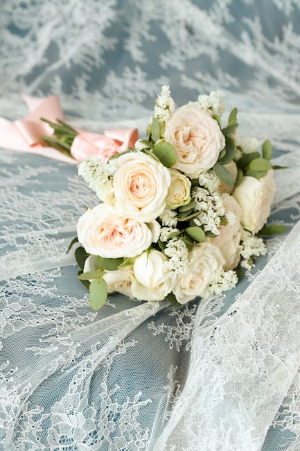 A delicate floral arrangement of fresh wedding bouquet featuring pale pink and white roses, complemented by small white blossoms and green eucalyptus leaves, lying on a soft blue lace fabric with intr