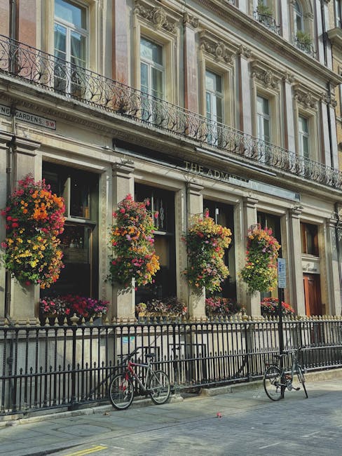 A vibrant floral arrangement featuring a mix of pink, orange, yellow, and red flowers, including petunias and geraniums, hanging in lush, overflowing baskets outside a historic building in Westminster