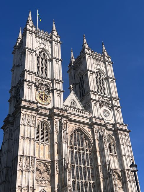 The image depicts Westminster Abbey's two prominent Gothic-style towers made of light-colored stone, featuring intricate detailing, arched windows, and clock faces. The sky above is clear and bright b